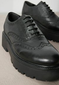 Chaussures en cuir noir à lacets avec des détails brogues, tige texturée et semelle plateforme épaisse avec un bord dentelé.