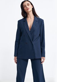 Granatowa marynarka dwurzędowa w pionowe paski, z klapami i dwiema czarnymi guzikami. W komplecie z dopasowanymi spodniami o szerokich nogawkach.