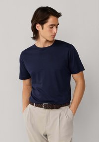 Joven con cabello oscuro que lleva una camiseta de manga corta azul marino metida en pantalones beige, de pie con las manos en los bolsillos y mirando hacia la izquierda.