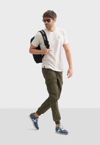 Camiseta de color claro, pantalones cargo oliva con bolsillos y zapatillas azules. Una mochila negra y gafas de sol completan el look casual.