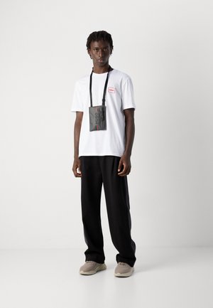 Ung mann iført en hvit t-skjorte, sorte bukser med sidelister, beige sneakers og en svart Hugo crossbody-veske som står mot en hvit bakgrunn.