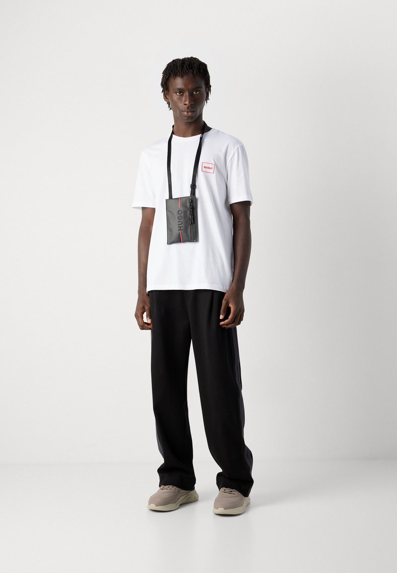 Ung mann iført en hvit t-skjorte, sorte bukser med sidelister, beige sneakers og en svart Hugo crossbody-veske som står mot en hvit bakgrunn.