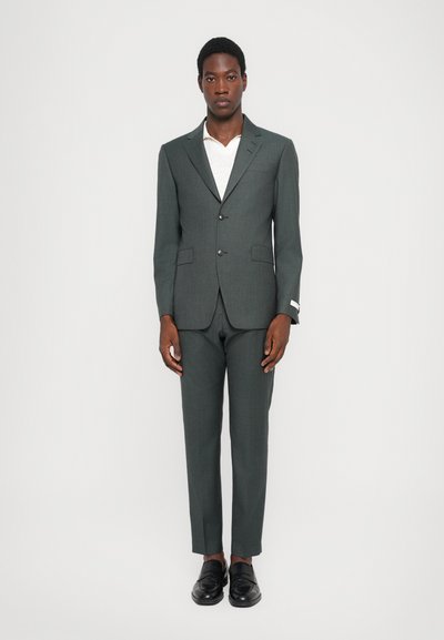 Completo grigio scuro con blazer strutturato, rever a V e pantaloni slim-fit. Abbinato a una camicia bianca e mocassini neri.