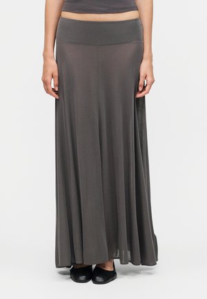 Žena nosí tmavošedú plynúcu maxi sukňu s vysokým pásom v kombinácii s čiernymi plochými topánkami, stojaca pred jednofarebným pozadím.