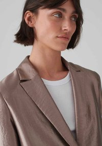 Metallisch taupefarbener Blazer mit Revers, strukturiertem Stoff und lockerer Passform, über einem weißen gerippten Shirt geschichtet.