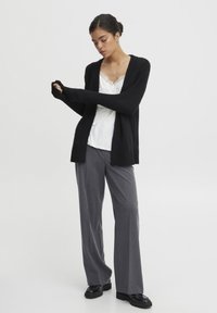 Svart långärmad cardigan med öppen framsida, ihopparad med en vit topp med spetsdetaljer och grå vida byxor. Svarta skor fulländar looken.