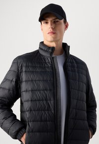 Young man wearing a black quilted jacket over a light grey shirt and a black baseball cap, standing with hands in pockets.