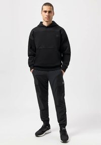 Sudadera negra con detalles de malla, bolsillos frontales y capucha con cordón, combinada con pantalones cargo oscuros que tienen bolsillos laterales y puños elásticos.