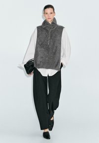 Gilet grigio e peloso con colletto alto e zip frontale, indossato sopra una camicia bianca a righe, abbinato a pantaloni neri a gamba larga e scarpe slip-on.