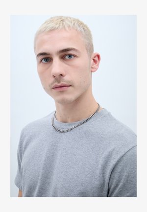 Jeune homme aux cheveux courts décolorés, aux yeux bleus, portant un t-shirt gris et une chaîne en argent, regardant droit devant.