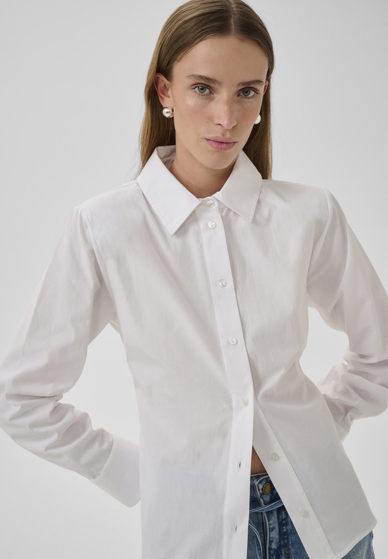 Jeune femme aux cheveux bruns raides, portant des boucles d'oreilles en perle, une chemise blanche boutonnée et un jean bleu, sur un fond clair uni.