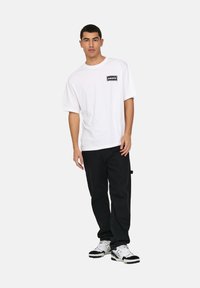 White short-sleeve t-shirt with a black rectangular "oasis" logo on the chest, paired with black pants and white sneakers with black accents.