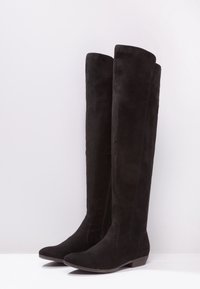 Botas de gamuza negra sobre la rodilla con tacón plano, que presentan una textura suave y un diseño sin costuras. Detalle de cremallera en un lado.