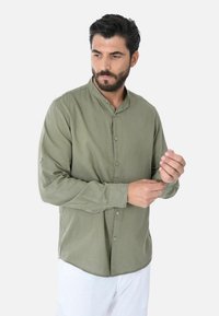 Camicia verde oliva a maniche lunghe con colletto a band, chiusura con bottoni e maniche arrotolabili, realizzata in un tessuto leggero con una consistenza liscia.