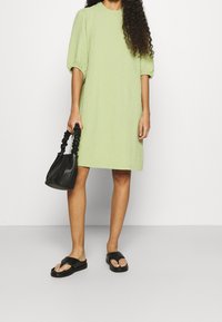 Robe courte à manches courtes, texturée et vert lime, avec une coupe décontractée, associée à un sac à main noir froncé et des sandales plates noires.