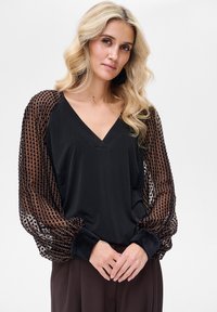 Czarny top z dekoltem w serek z przezroczystymi brązowymi, wzorzystymi rękawami i czarnymi aksamitnymi mankietami. Rękawy mają wzór w groszki, a tkanina wydaje się miękka.