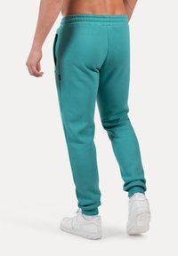 Pantalones de chándal verde azulado hechos de una tela suave, con piernas ajustadas, cintura elástica, bolsillos laterales y puños acanalados. Combinados con zapatillas blancas.