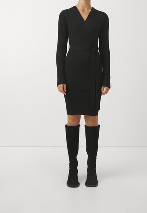 Kobieta ubrana w czarną prążkowaną dzianinową sukienkę kopertową z długimi rękawami oraz czarne zamszowe kozaki sięgające do kolan, na jednolitym białym tle.