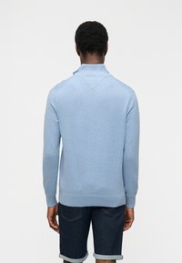 Άνδρας που φοράει ανοιχτό μπλε πουλόβερ με μακριά μανίκια και σκούρα τζιν σορτς με γυρισμένα μπατζάκια, στέκεται γυρισμένος με την πλάτη σε λευκό απλό φόντο.