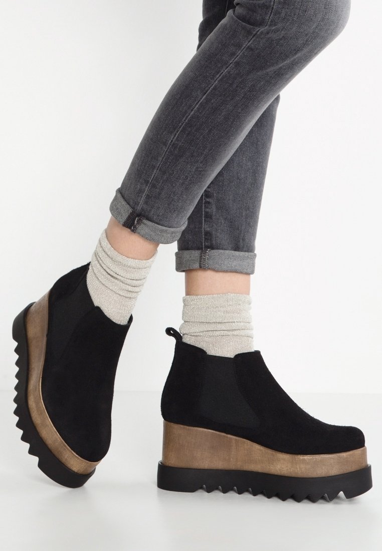 Bottes de cheville en suède noir avec une semelle noire striée et un intercalaire doré métallique. Portées avec un jean gris retroussé et des chaussettes crème.