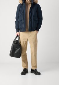 Navy pufferjack met een met bont gevoerde capuchon, lichtbruine coltrui, beige chino's, zwarte leren tas en zwarte veterschoenen.