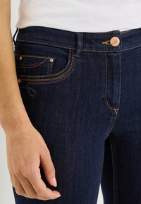 Jean en denim bleu foncé à coupe ajustée, avec des coutures orange, un bouton en cuivre, et des poches avant ornées de coutures décoratives.