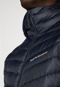 Námořnická puffer bunda s vysokým límcem, horizontálním prošíváním a zipem. Obsahuje logo "PeakPerformance" bílé výšivky.