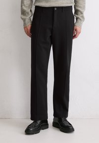 Pantalons noirs avec une coupe droite, des plis avant, une fermeture à boutons et une texture lisse. Associés à des chaussures noires à semelle texturée.