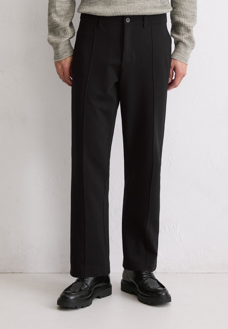 Pantalons noirs avec une coupe droite, des plis avant, une fermeture à boutons et une texture lisse. Associés à des chaussures noires à semelle texturée.
