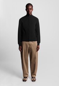 Sort zip-up jakke med høj krave, tekstureret stof, sidelommer; parret med beige bukser og mønstrede brune sneakers.