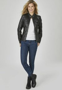 Schwarze Lederjacke mit Kragen, zwei Fronttaschen, kombiniert mit einem weißen T-Shirt, dunkelblauer Jeans und schwarzen Schnürstiefeln.