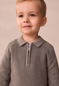 Brauner Reißverschluss-Pullover mit strukturierter Strickmusterung, geripptem Kragen und kurzem Reißverschluss, getragen von einem Kind vor neutralem Hintergrund.