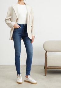 Beige linnblazer över en vit topp, matchad med blå skinny jeans och vita sneakers med bruna sulor, mot en neutral bakgrund.