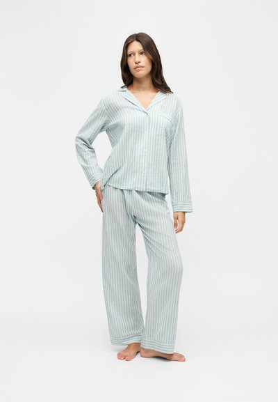 Femme portant un pyjama rayé vertical bleu clair et blanc à manches longues avec chemise boutonnée et pantalon ample, debout pieds nus sur fond blanc.