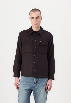Veste marron foncé en matériau doux, avec deux poches poitrine, des boutons argentés et un accent jaune sur le côté gauche. Jean bleu.