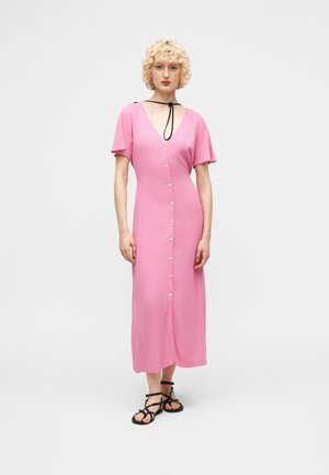 Γυναίκα με σγουρά ξανθά μαλλιά, φοράει ένα μακρύ ροζ φόρεμα με κουμπιά και μαύρα σανδάλια με λουράκια, στέκεται μπροστά σε ένα απλό φόντο.