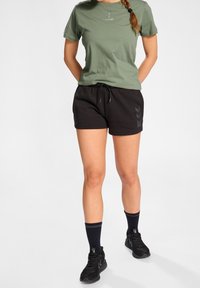 T-shirt in cotone verde oliva con piccolo logo, abbinato a pantaloni corti neri con cordoncino e motivi a chevron sui lati. Scarpe da ginnastica nere e calze.