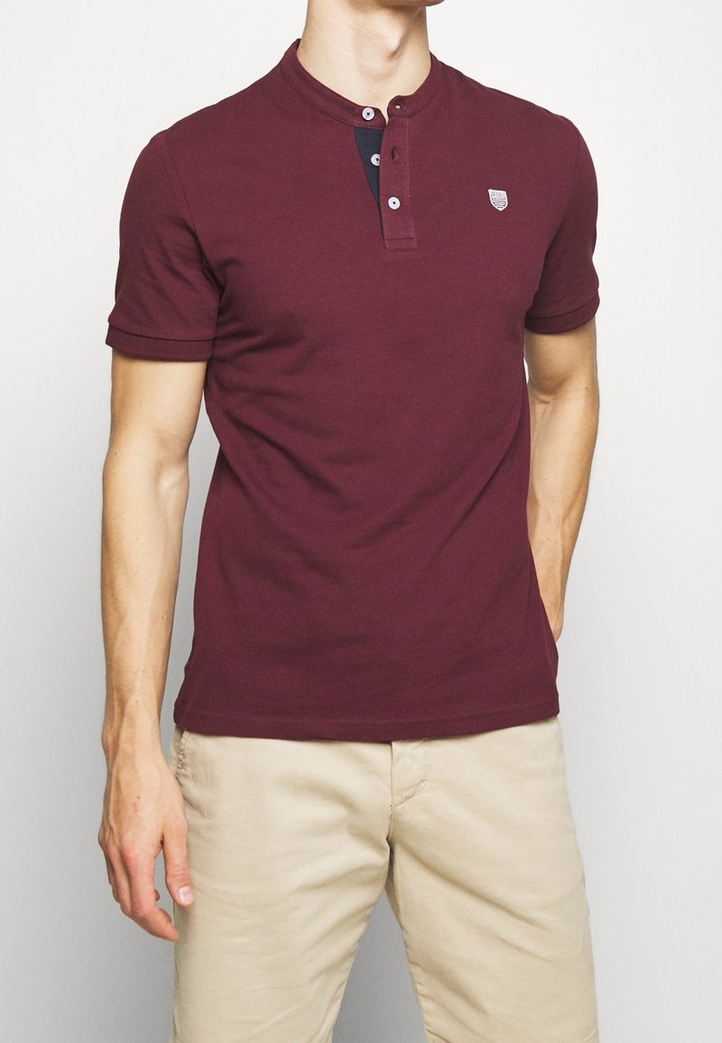 Polo bordeaux à manches courtes, col henley et patte de boutonnage à trois boutons. Il présente un petit logo sur la poitrine gauche et une texture lisse.