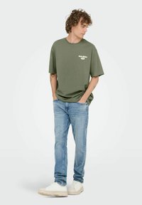 Olijfgroene katoenen t-shirt met korte mouwen, voorzien van witte "Solo Rider" tekst, gecombineerd met lichtblauwe jeans en witte sneakers.