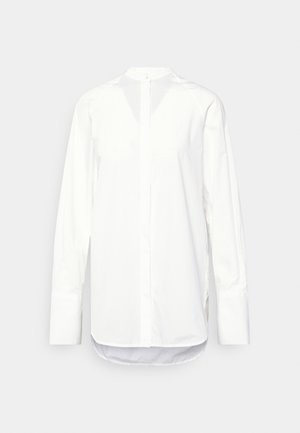 Chemise blanche à manches longues avec patte de boutonnage cachée, encolure ronde, poignets allongés et ourlet arrondi sur un fond neutre.