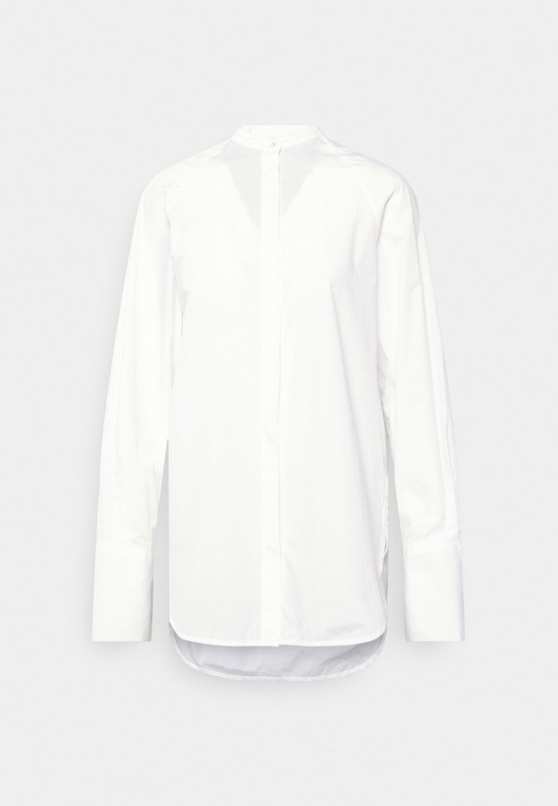 Chemise blanche à manches longues avec patte de boutonnage cachée, encolure ronde, poignets allongés et ourlet arrondi sur un fond neutre.