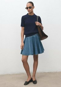 Femme portant des lunettes de soleil noires, un pull court bleu marine à manches courtes, une jupe en denim bleue, des ballerines noires et un sac à main marron sur l'épaule, contre un mur blanc.