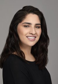 Portret van een vrouw met lang, donker, golvend haar, die glimlacht. Ze draagt een zwarte top tegen een neutrale achtergrond, met natuurlijke makeup en heldere tanden.