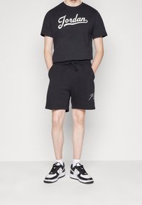 Černé bavlněné tričko s logem "Jordan" v bílé barvě, spárované s černými teplákovými šortkami s tkaničkou a bílým grafickým akcentem. Černobílé tenisky.