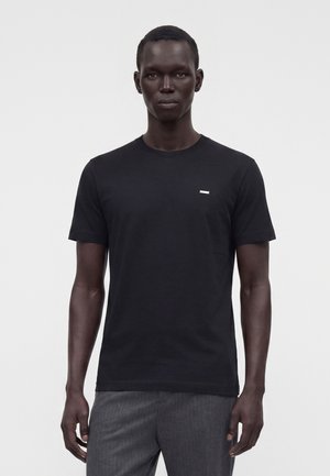 Mand stående iført en ensfarvet sort t-shirt med korte ærmer og grå bukser mod en hvid baggrund.