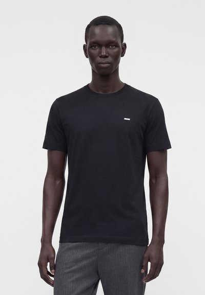 Homme debout portant un t-shirt noir uni à manches courtes et un pantalon gris sur un fond blanc.
