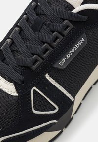 Marinblå sneakers med ovandel i mocka och textil, svarta snören, vita detaljer och en präglad Emporio Armani-logotyp på sidan.