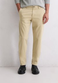 Khakifarbenes Chino mit glattem Baumwollstoff, ausgestattet mit Fronttaschen und einem Knopfverschluss, kombiniert mit schwarzen, glänzenden Schuhen.