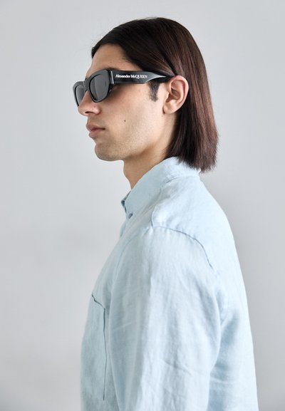 Occhiali da sole neri con montatura spessa e lenti scure. Il nome del marchio "Alexander McQUEEN" è stampato sul lato. Indossati con una camicia azzurra.