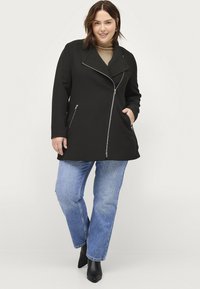 Zwarte op maat gemaakte jas met een dubbele kraag en zilveren ritsen, met twee zijzakken, gecombineerd met lichtblauwe rechte jeans en zwarte laarzen.